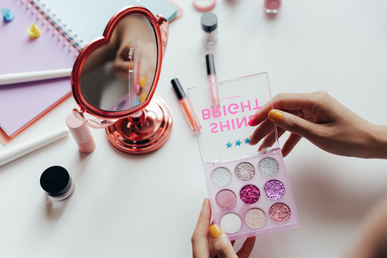 A vibrant eyeshadow palette with glittery colors held near a heart-shaped mirror.