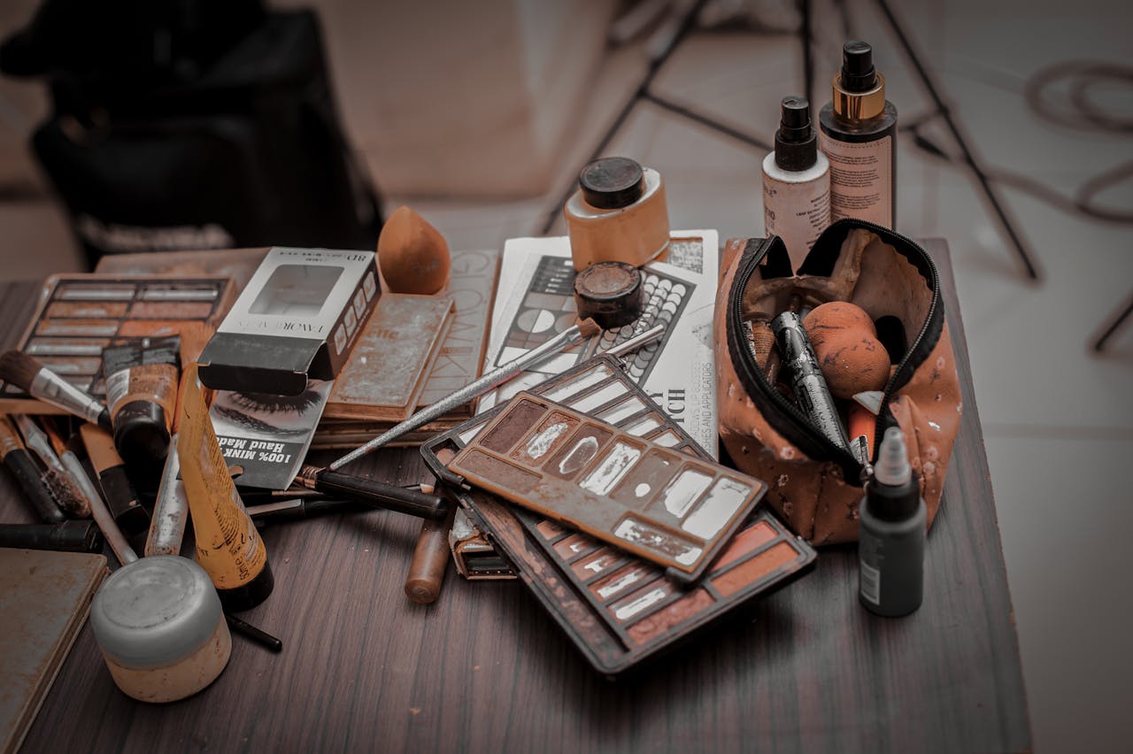 A collection of used makeup products, including brushes and palettes, on a wooden table indoors.