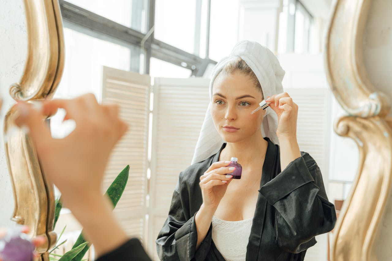 A woman in a towel and robe applies skincare serum with a dropper in front of a mirror.